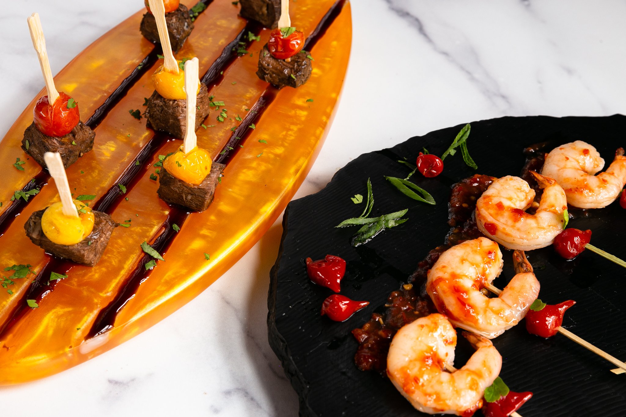  Sesame Shrimp Skewers are lined up and plated on a black serving dish next to Balsamic Beef and Roasted Tomato Skewers, arranged on a long, golden platter. The platters sit on a white marble surface. 