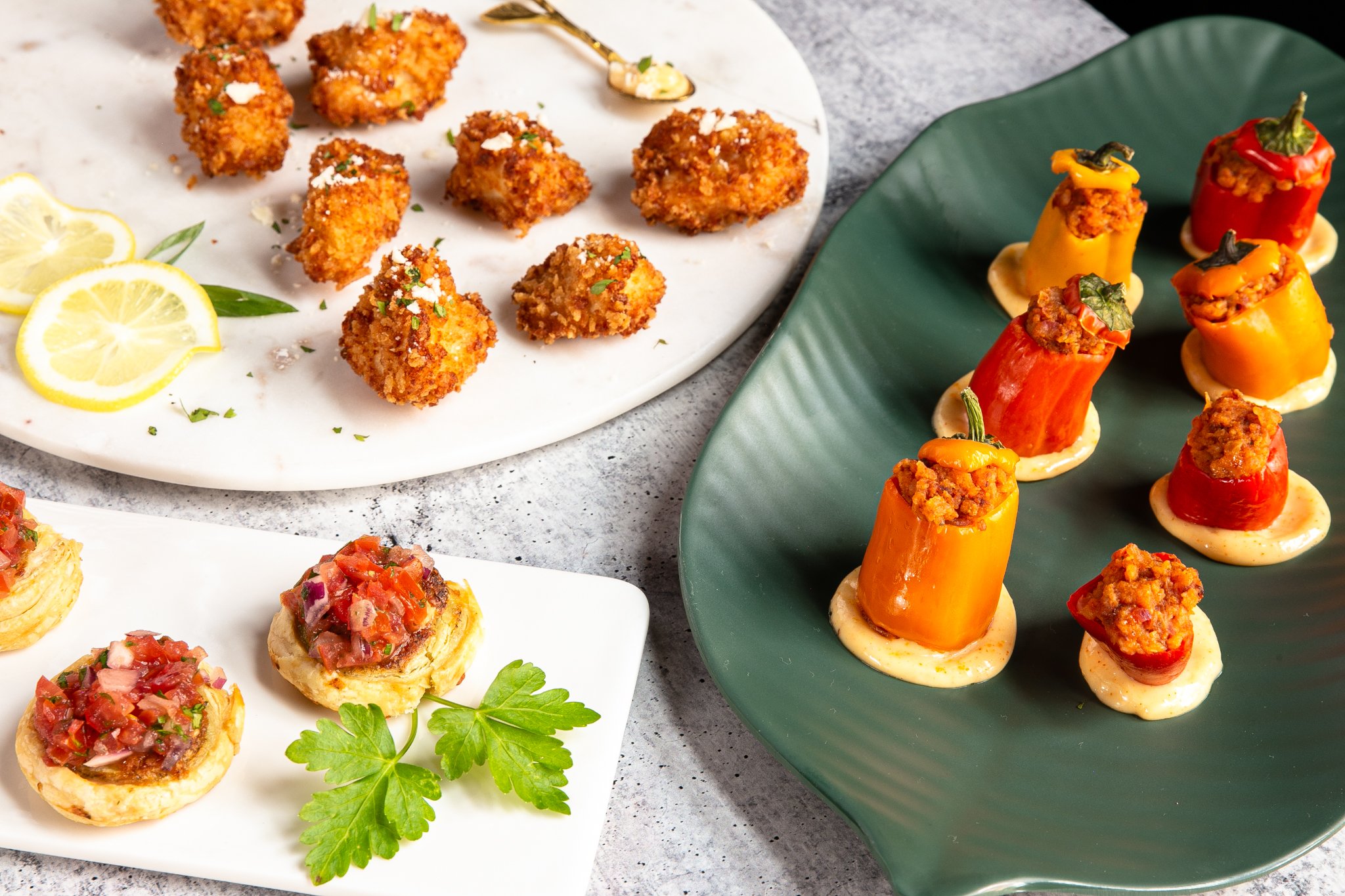  Platters of several different hors d’oeuvres, including Parmesan Herb Chicken Bites, Tomato Pesto Palmier, and Chorizo Stuffed Peppers, are placed next to each other on a light gray countertop. 