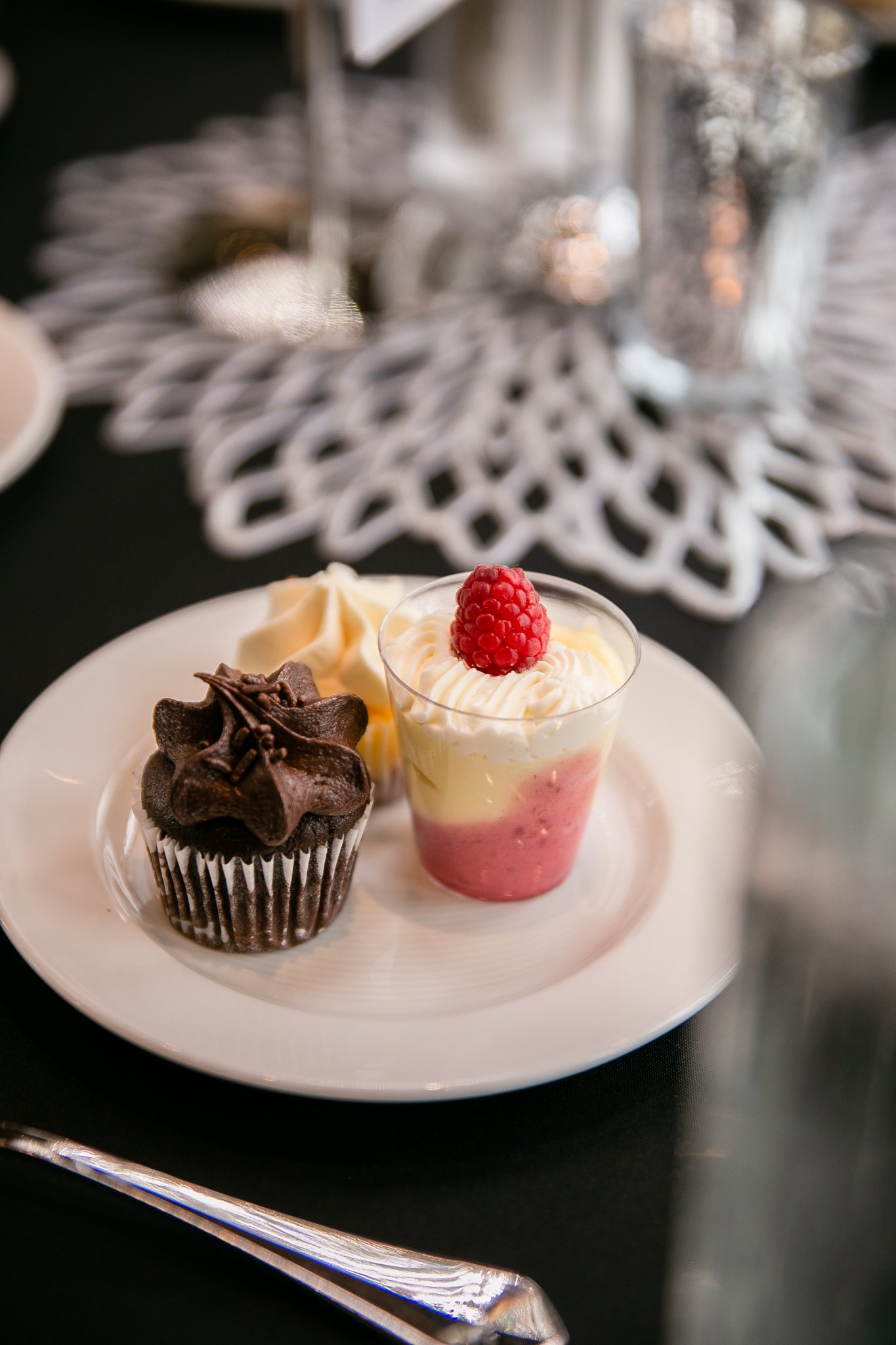  An arrangement of Signature Minature Trio desserts (mini chocolate cakes, mini cheesecake, and seasonal mousse) is plated for an event. A decorative doily is in the center of the table in the background. 