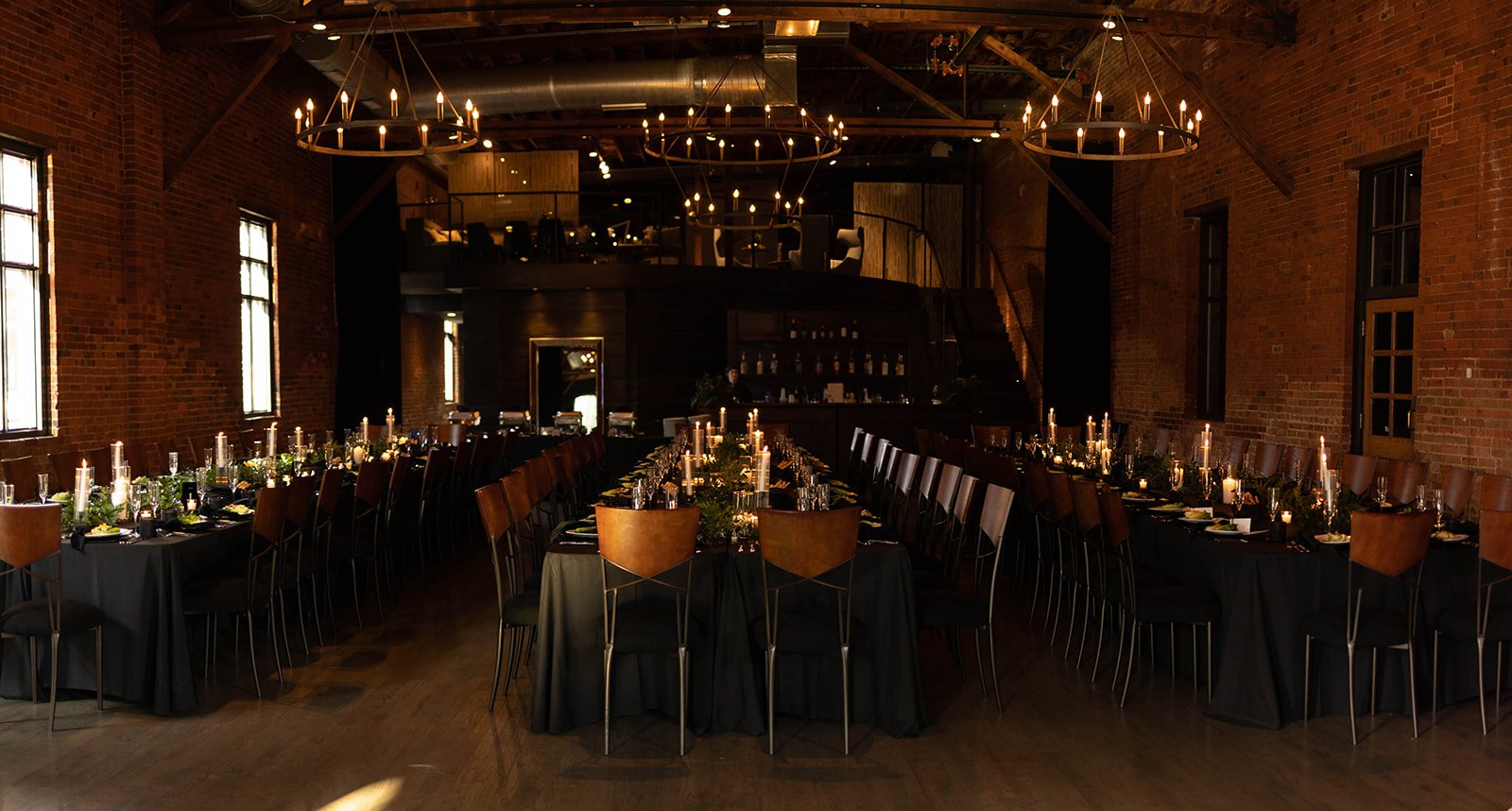  A moody, dimly lit reception hall with long tables, black tablecloths, and candlesticks down the center of the tables. Two-tiered rustic candelabra chandeliers hover over the room. 