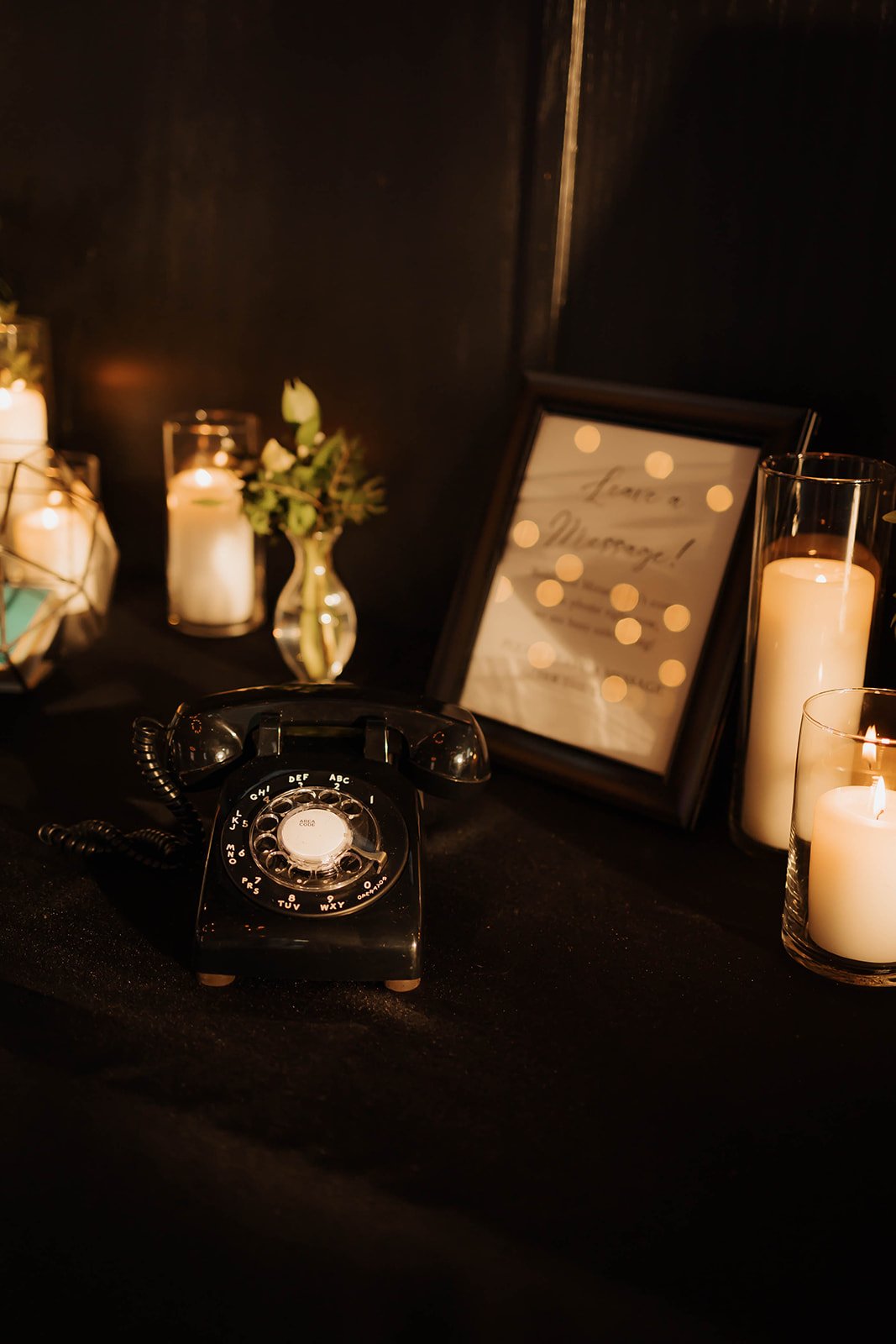  A black rotary phone sits on a table with a sign next to it that says “Leave A Message!”. White candles and a small vase of flowers frame the phone in the background.  