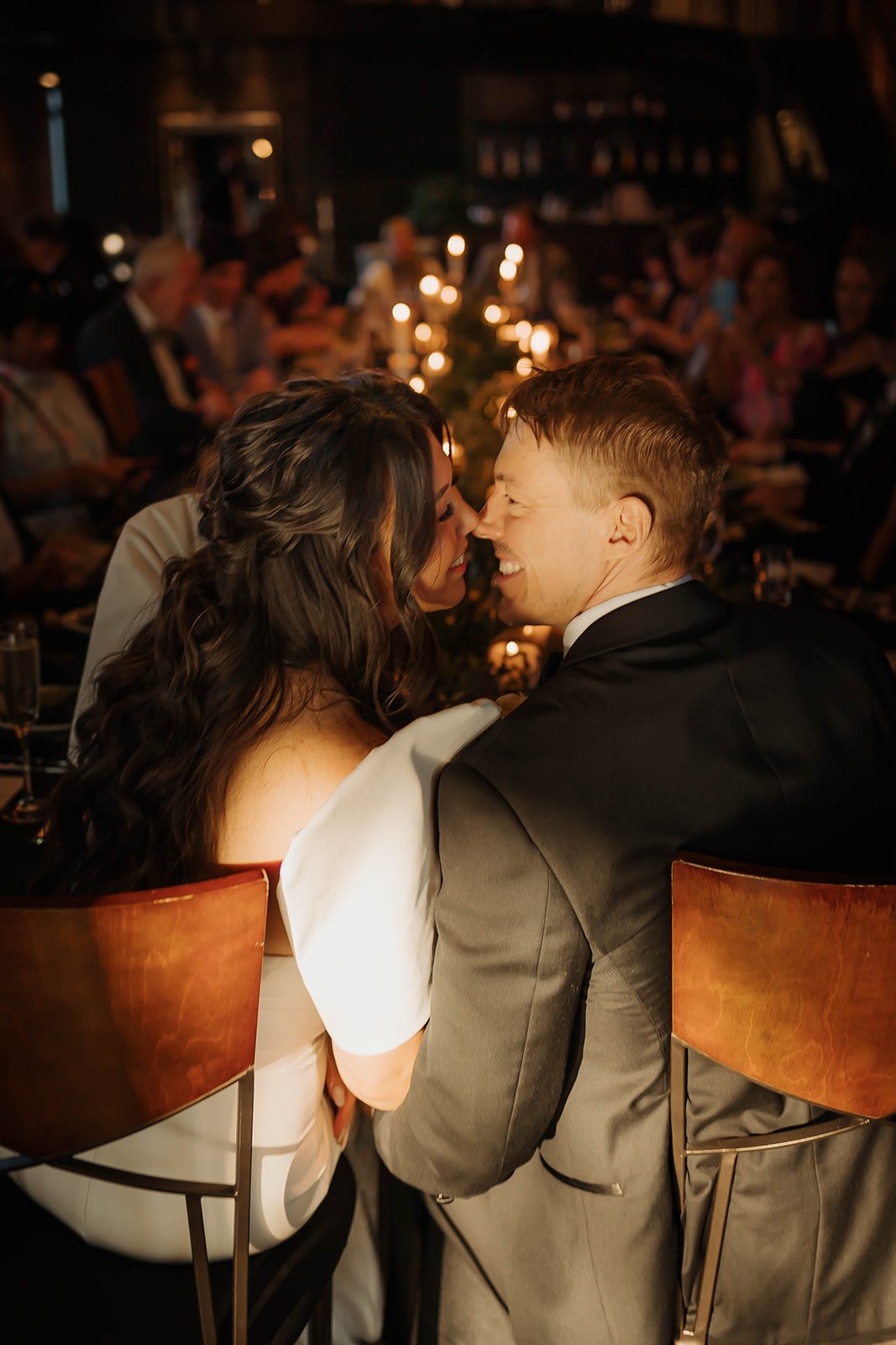 A bride and groom sit at the head of a long wedding reception table, leaning in to kiss each other as sunlight coming in the tall windows washes over them. 