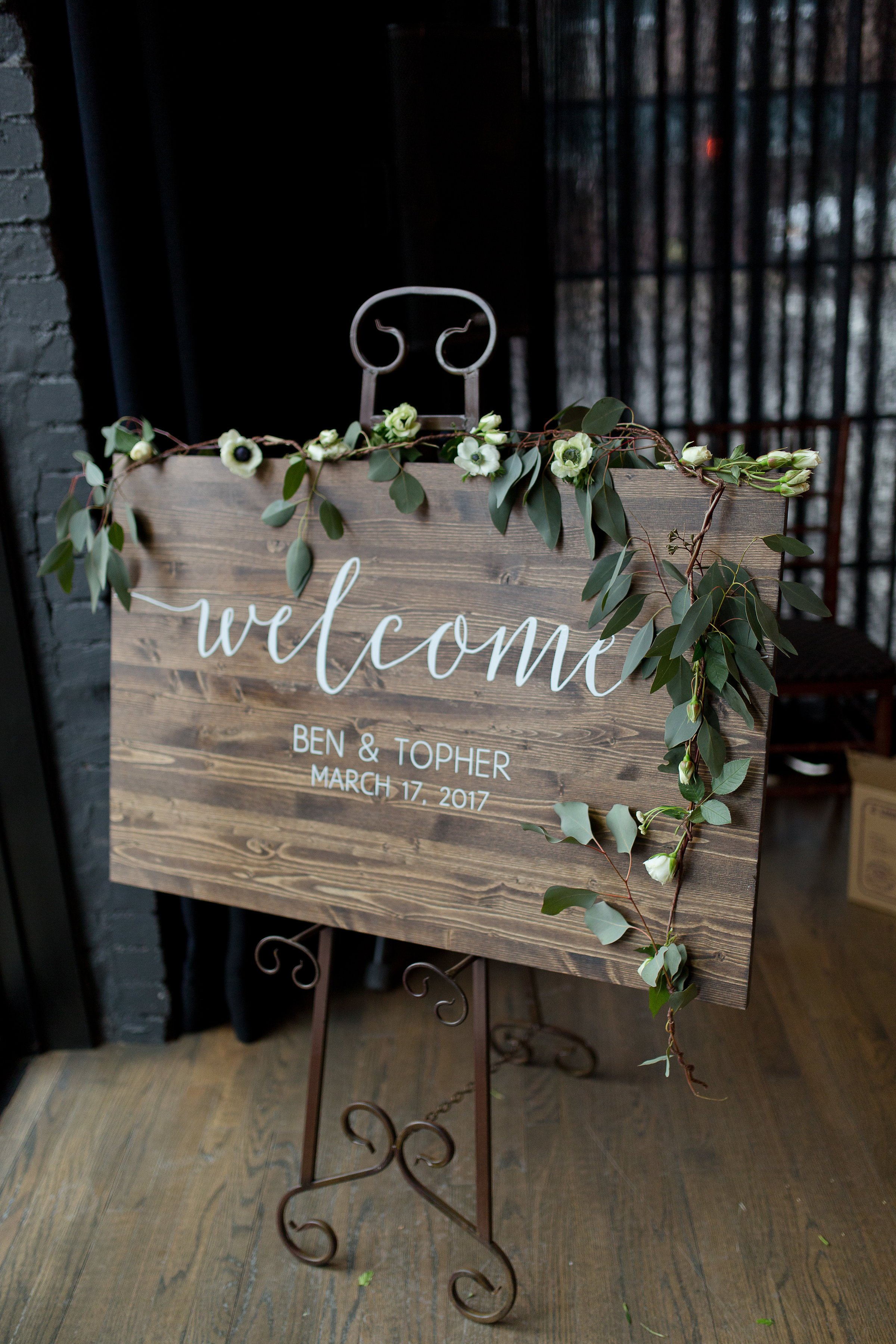  A welcome sign written on a wooden board and decorated with flowers at the entrance of a wedding reception. The sign reads “Welcome Ben &amp; Topher, March 17, 2017” 