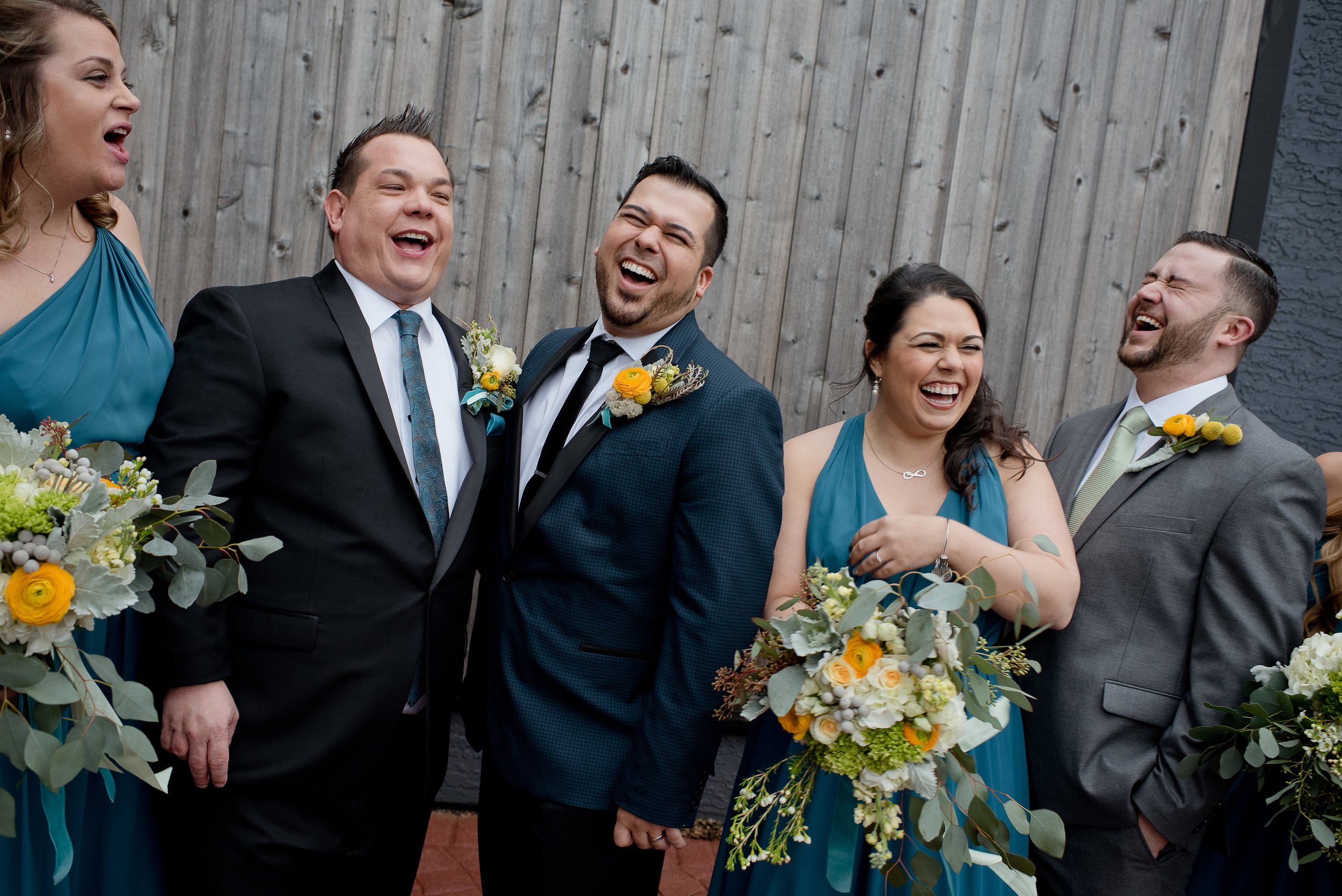  Two grooms stand together on the High Line Car House patio with their wedding party, all laughing together before their wedding ceremony. 