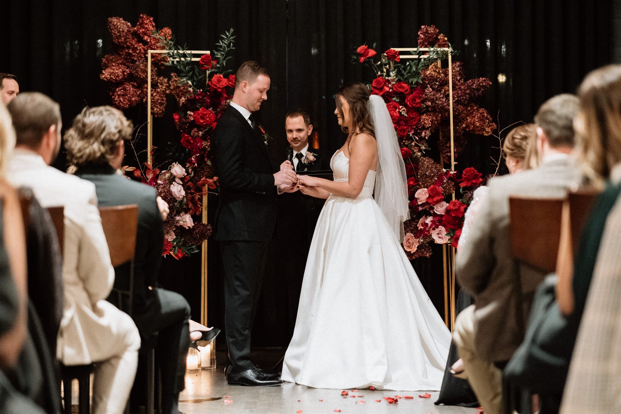  A couple stands at their wedding altar as a groom  a ring on a bride’s finger. Their altar is made of geometric gold flower stands that frame the couple as they take their vows. 
