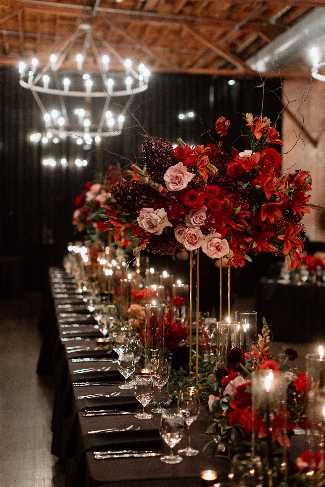  A view down a long black linen table set for a wedding reception dinner. Down the center of the table are tall candlesticks and small red floral centerpieces, interspersed with tall gold stands of large red and pink floral centerpieces. The table st