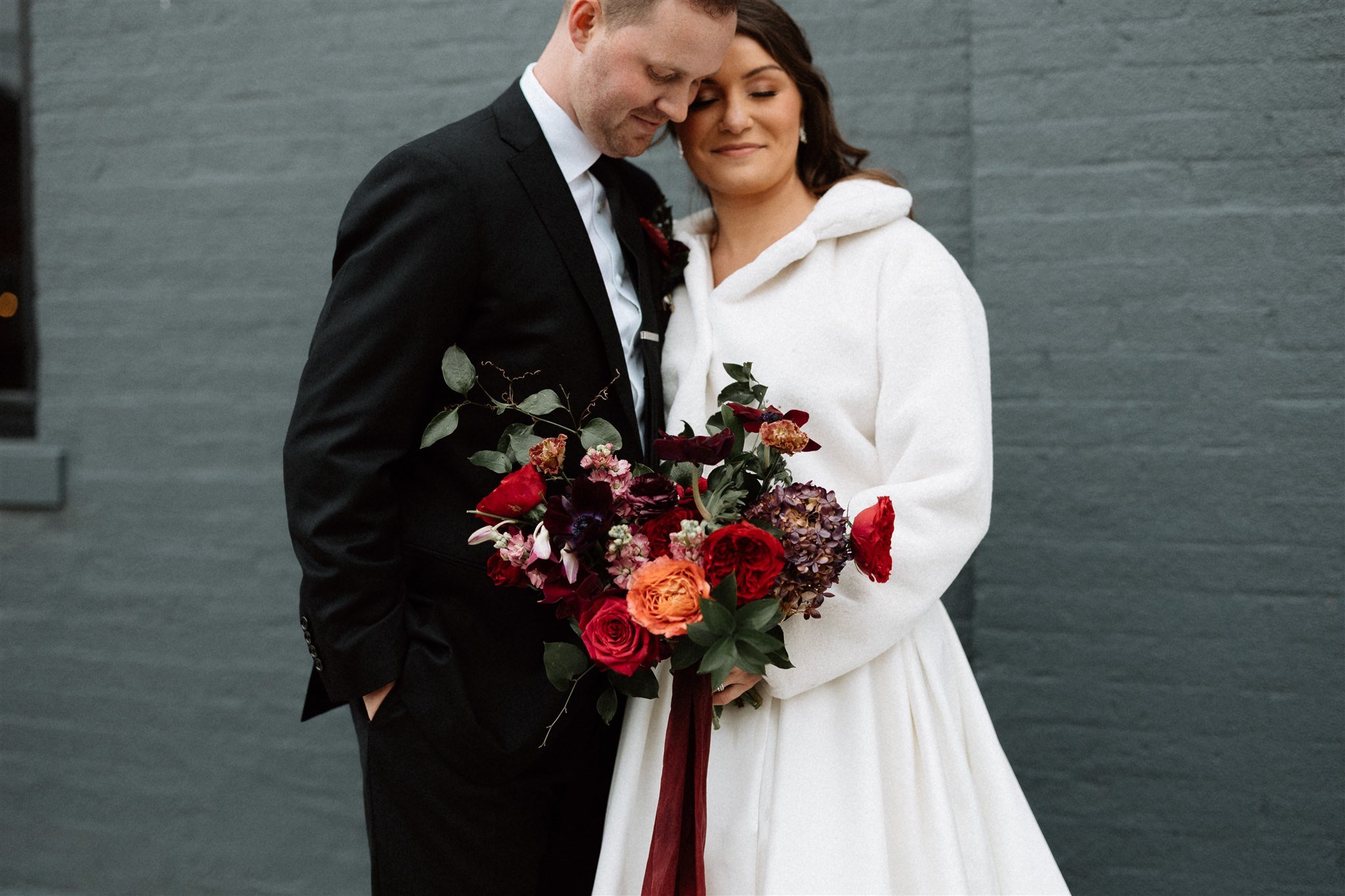  A couple lean in towards each other, smiling, outside a brick wedding venue. The bride is wearing a white furry coat and holding a large bouquet of dramatic red and orange flowers, accented by green leaves.  