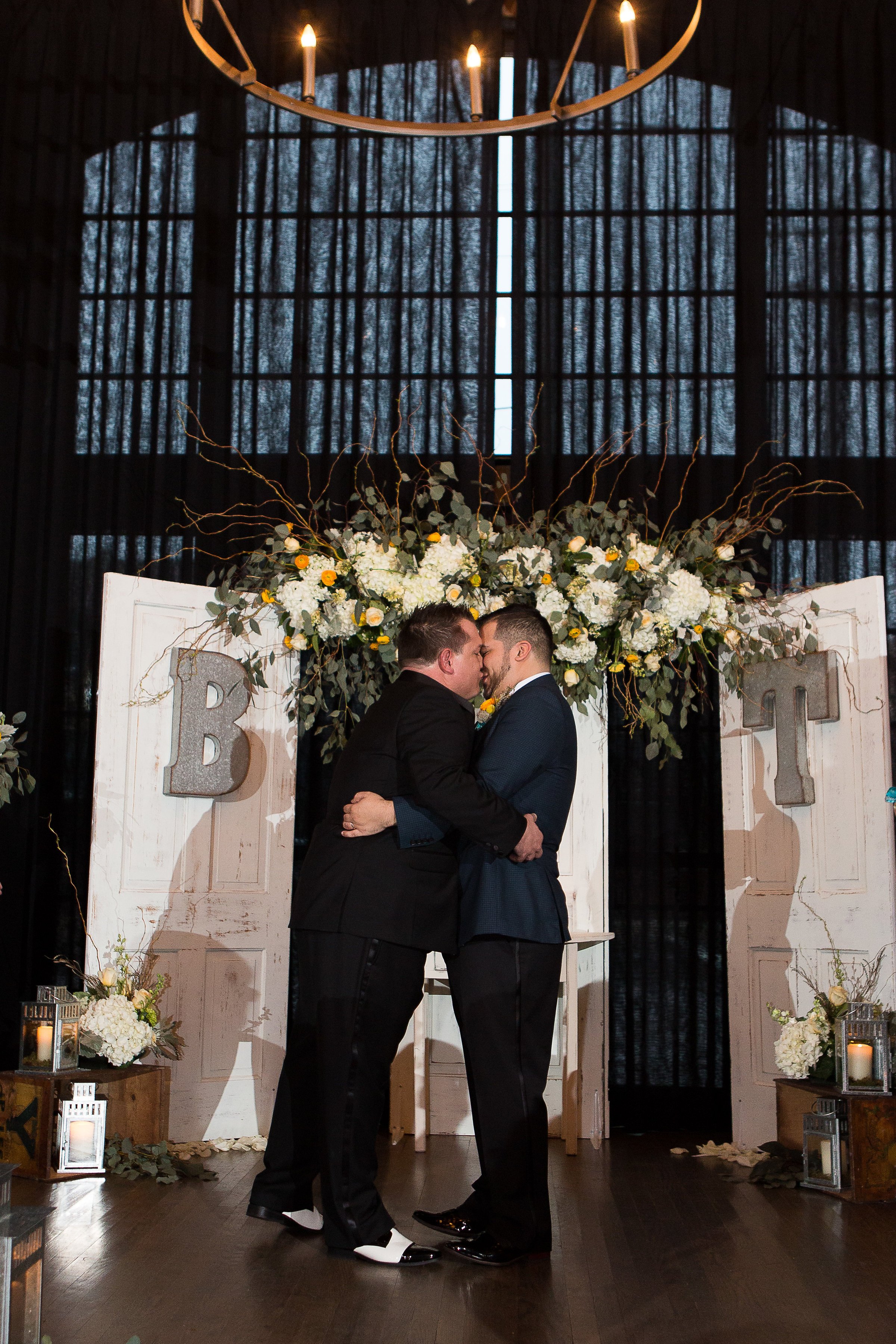  Two grooms embrace and kiss at their altar made up of doors and a beautiful floral tapestry within an industrial wedding venue.  