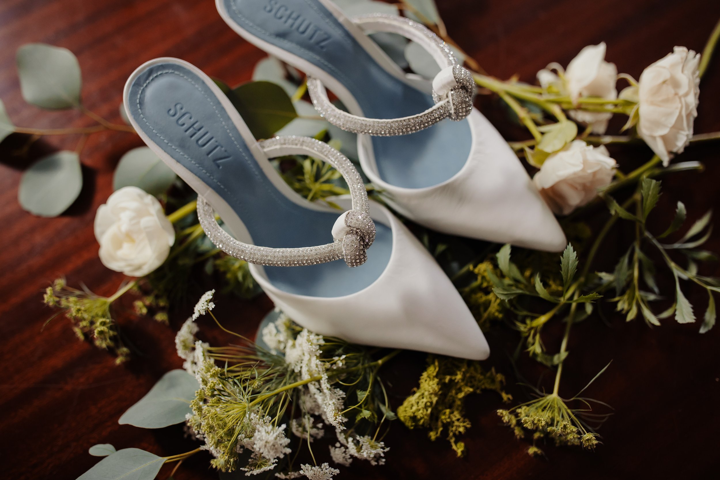  Closeup of a pair of bridal shoes next to some table florals. The shoes are white with light blue insoles. 