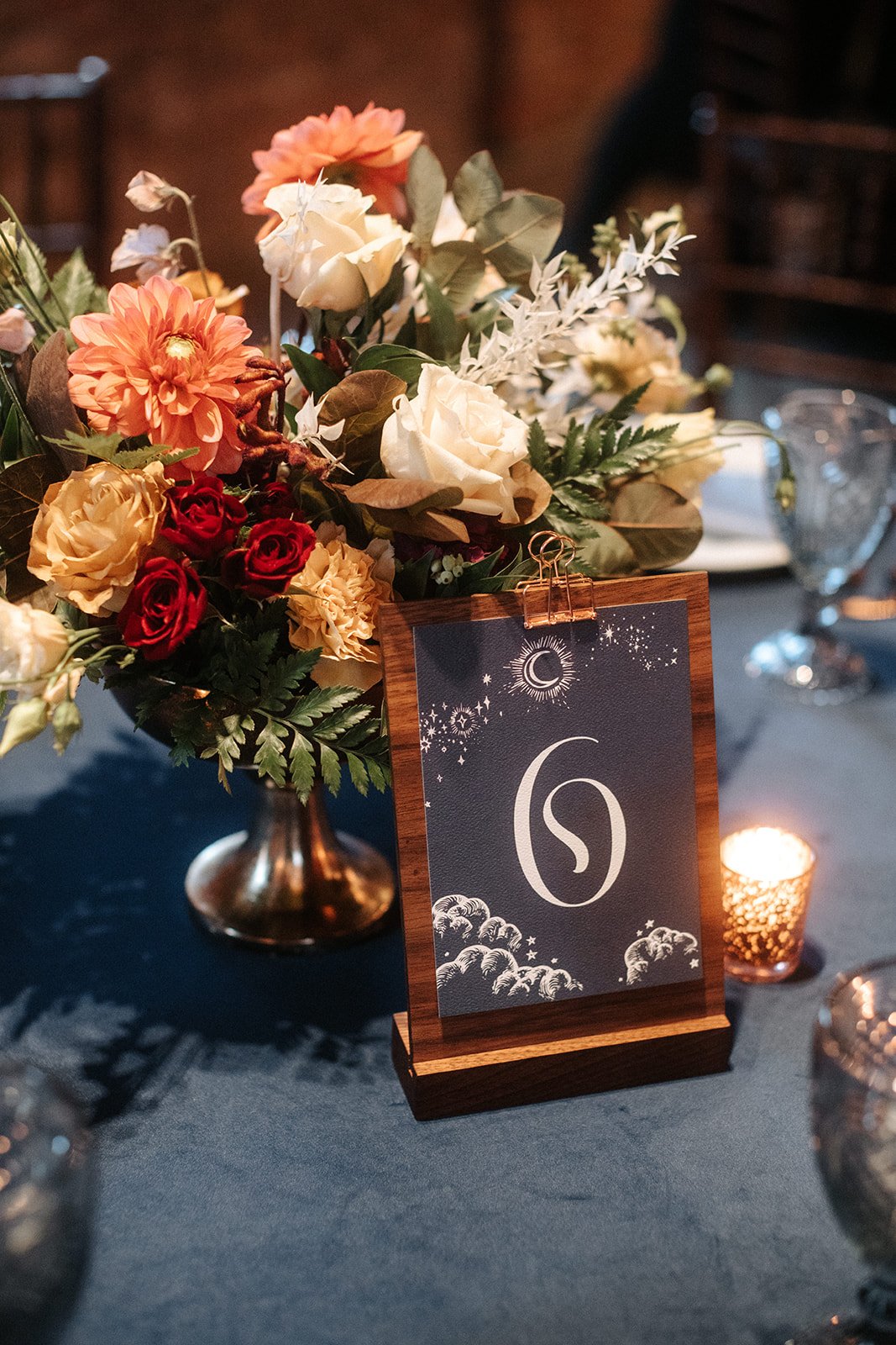  A table number designed in an ethereal style is mounted on a wood stand. Behind the number is a lush bouquet of florals in a short, silver flower stand.  