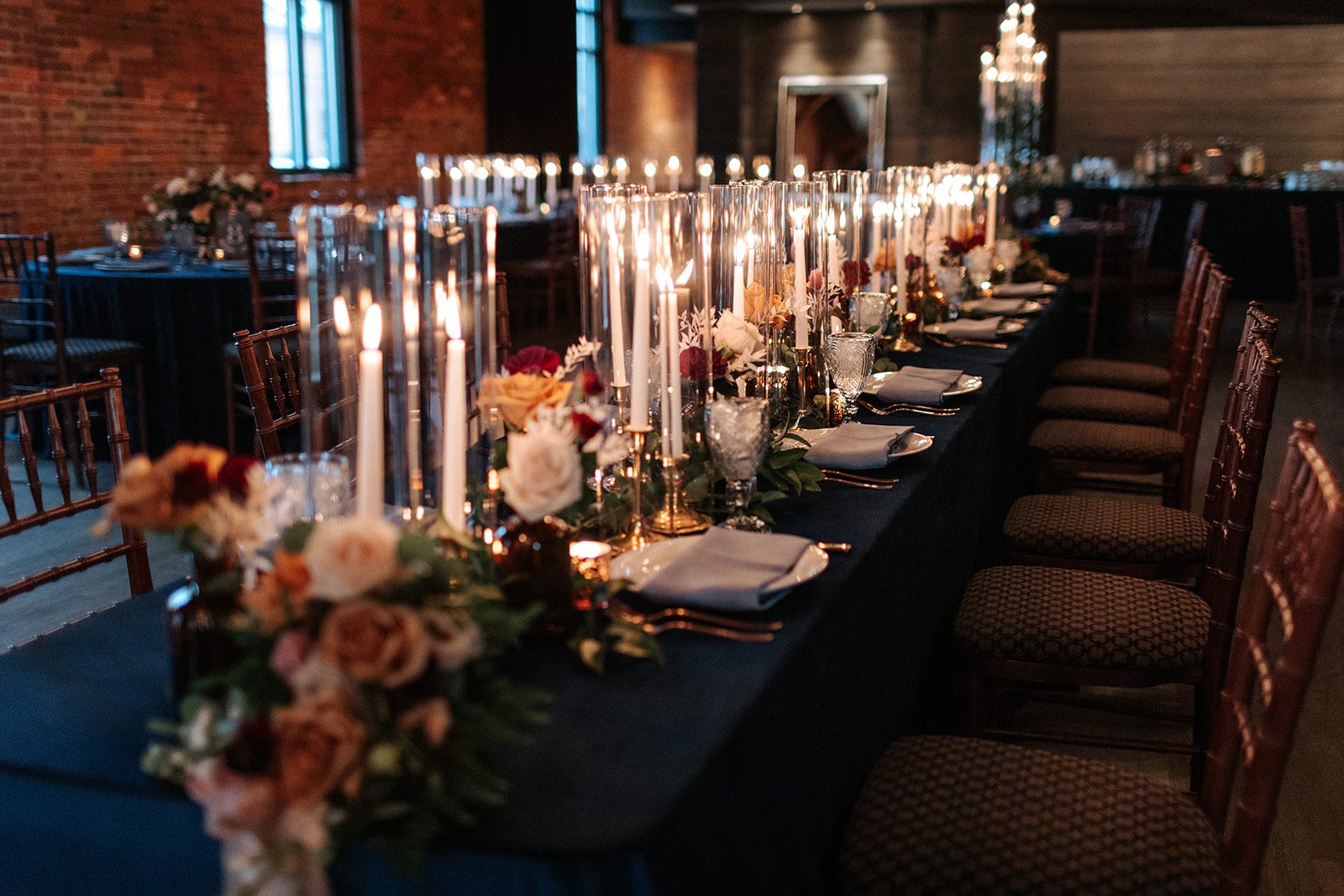  An industrial wedding venue in Columbus, Ohio, is filled with long tables decorated with small floral centerpieces and tall white candlesticks placed in glass hurricanes, all lit for the wedding reception dinner. 