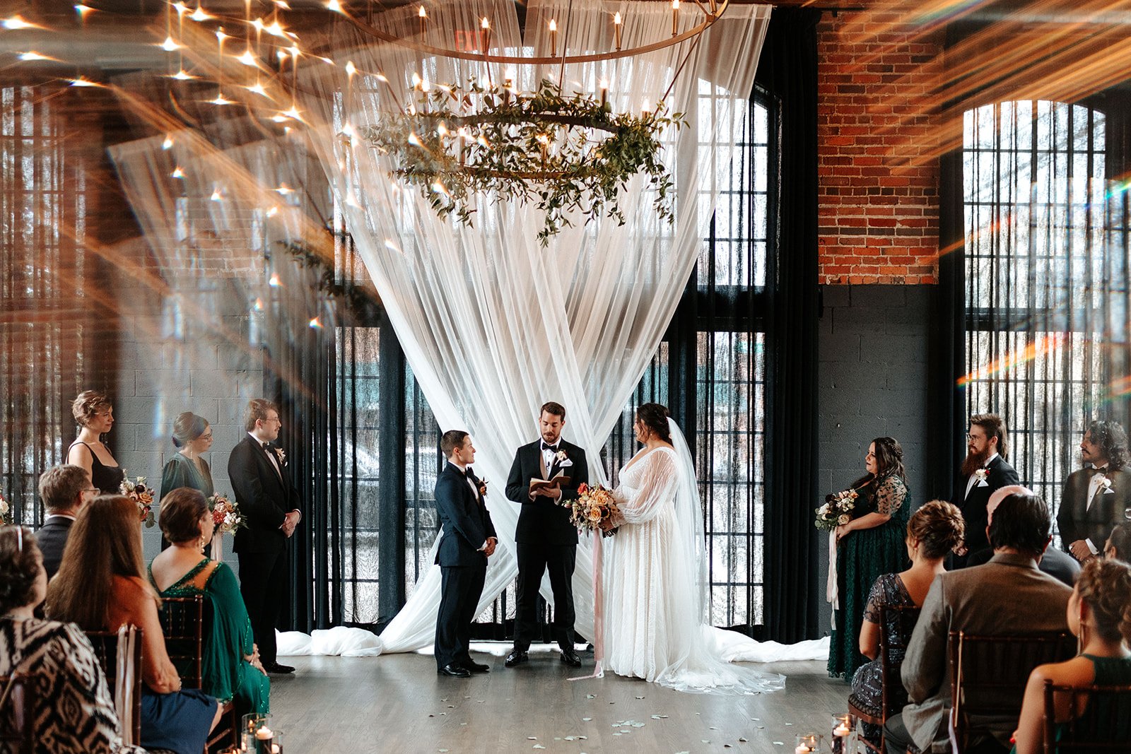  A couple stands at their altar with a sheer white tapestry backdrop behind them as they listen to their officiant. The two-tiered chandelier above them is decorated with greenery, and their wedding party watches the ceremony from either side.  