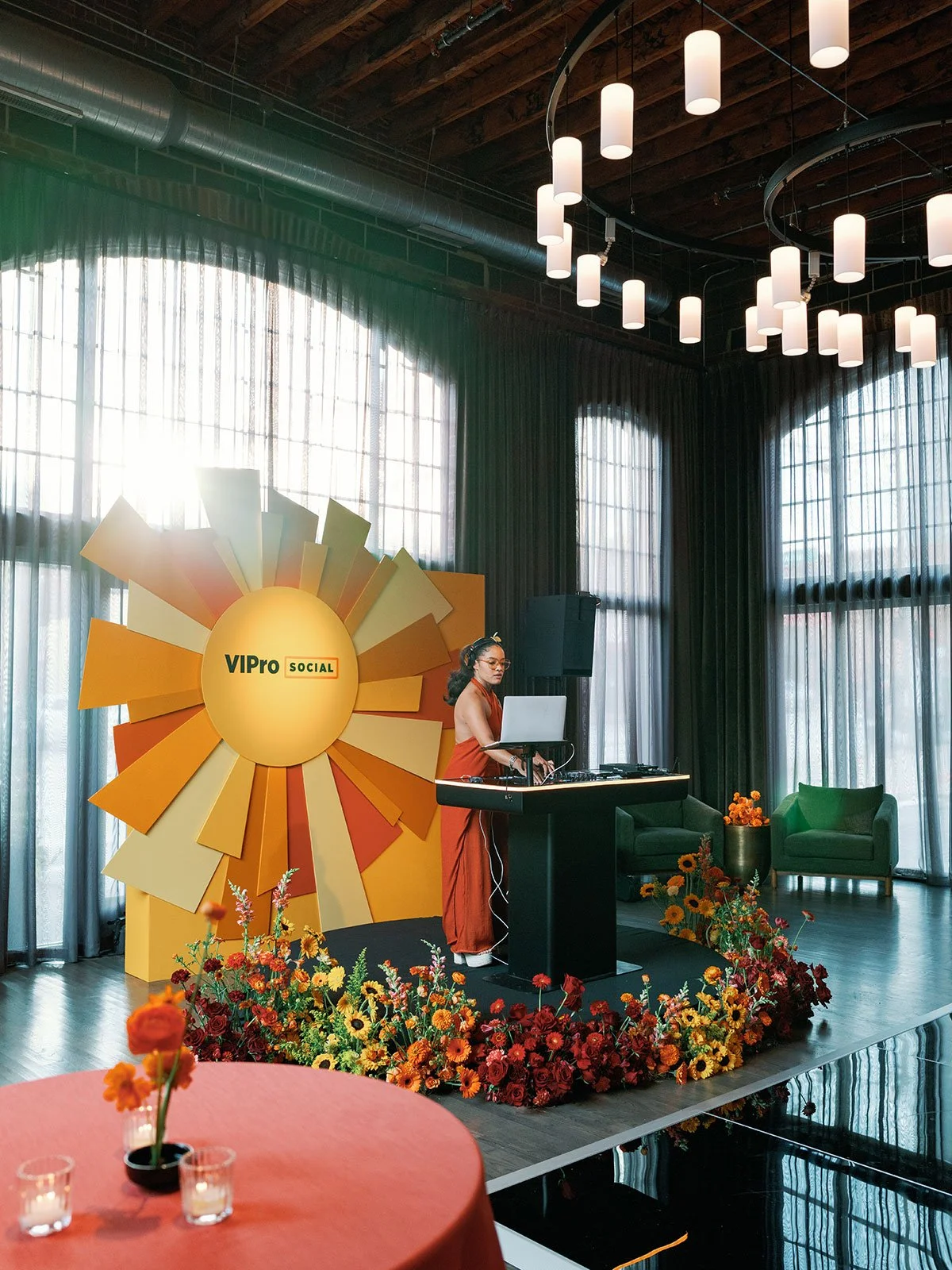 A DJ plays music from a small stage in the Grande Hall at High Line Car House in German Village. Behind them is a sturdy backdrop made of a cutout of a sun and its rays. The stage is decorated with red, orange, and yellow flowers.