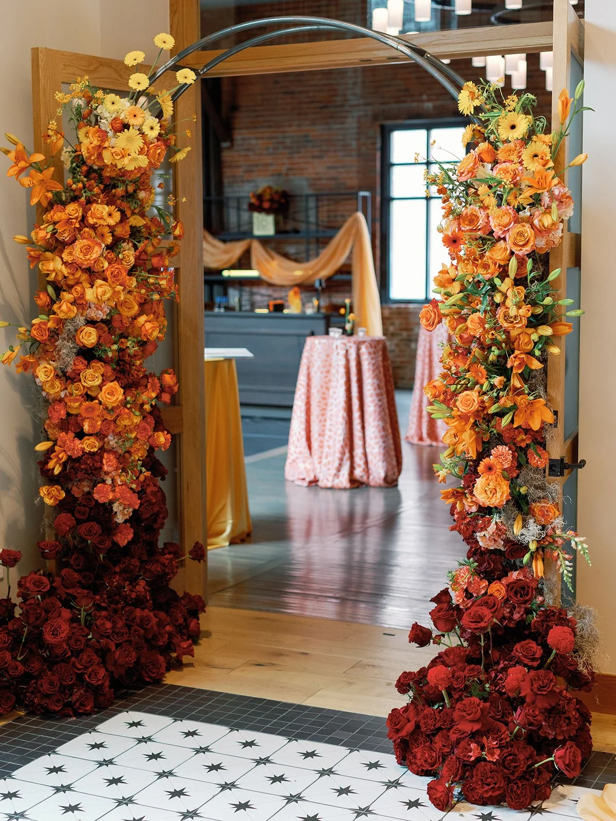 A metal arch stands at the entrance of a doorway between two event spaces at the industrial wedding venue, High Line Car House, decorated entirely in red, orange, and gold florals.