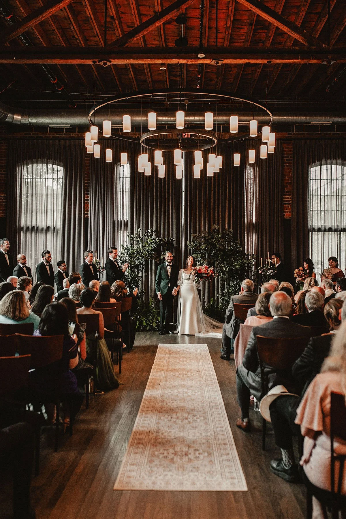A bride and groom stand at the end of an aisle at a downtown industrial event venue, smiling at their guests as they applaud. There are trees with dark green leaves behind them as their ceremony backdrop.