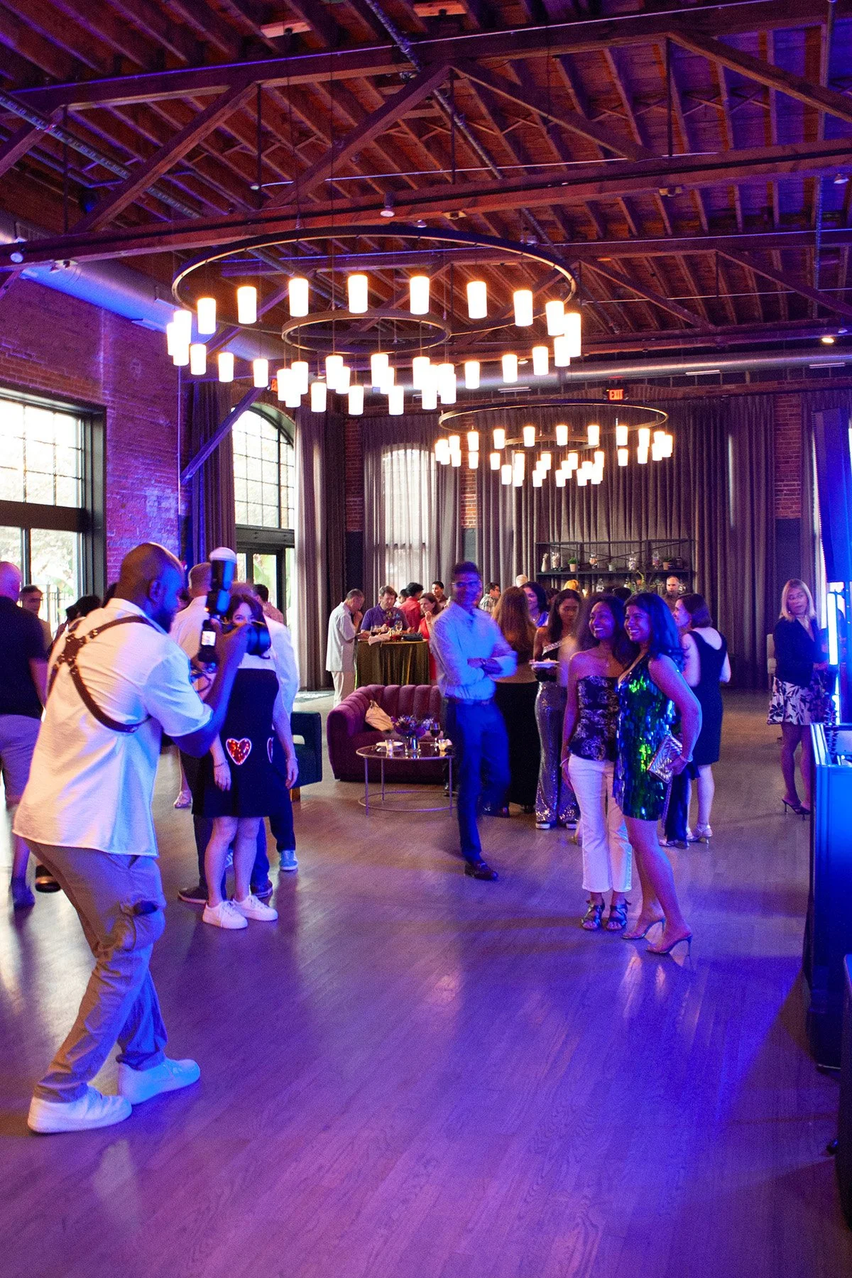 A photographer takes a picture of guests at a birthday party hosted at a spacious venue in downtown Columbus, Ohio. There are large contemporary chandeliers above them, ands purple uplighting accents the room.
