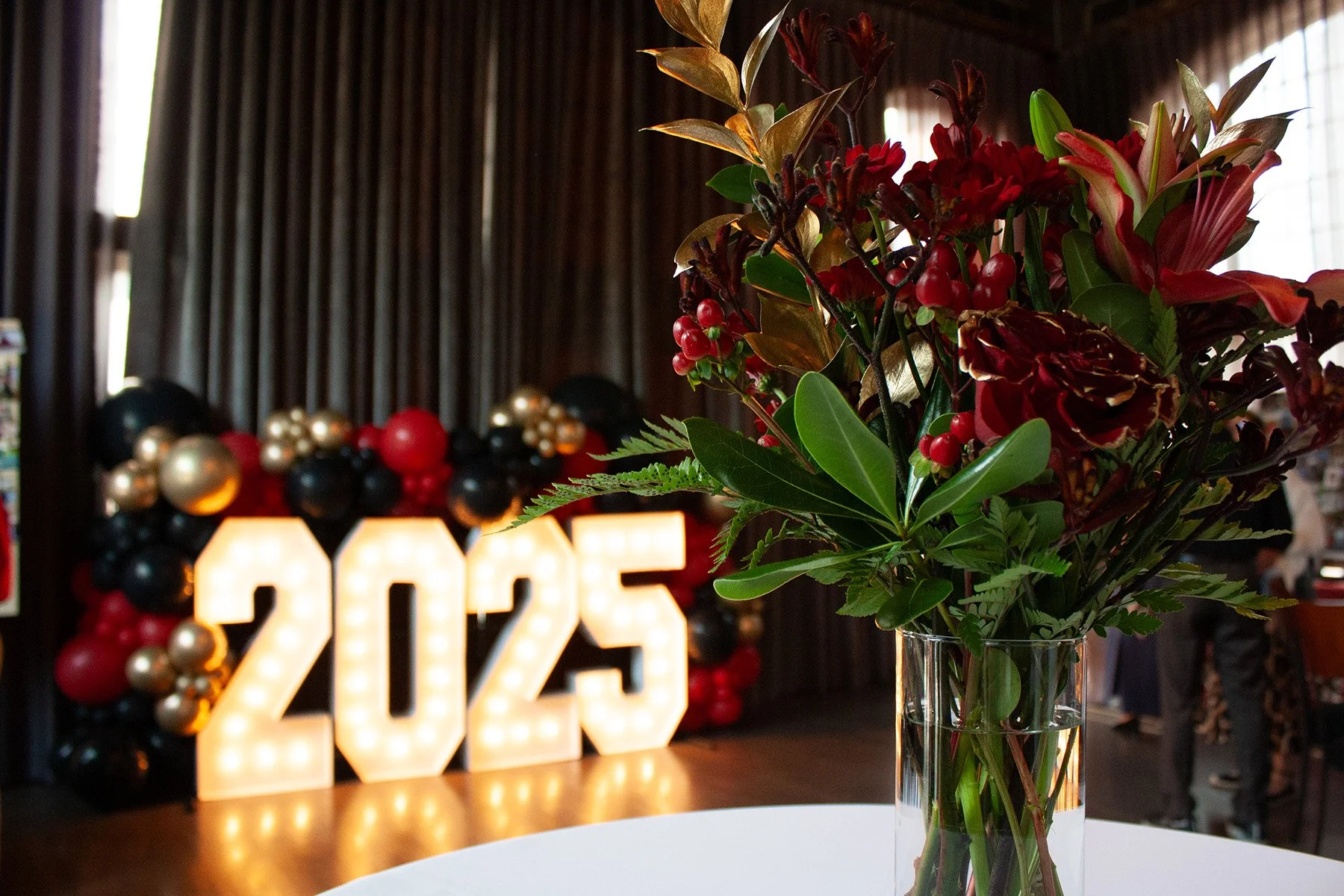 A festive, red and gold floral arrangement is in the middle of a tallboy table at a graduation party. A large marquee sign with balloon decor around it is in the background.