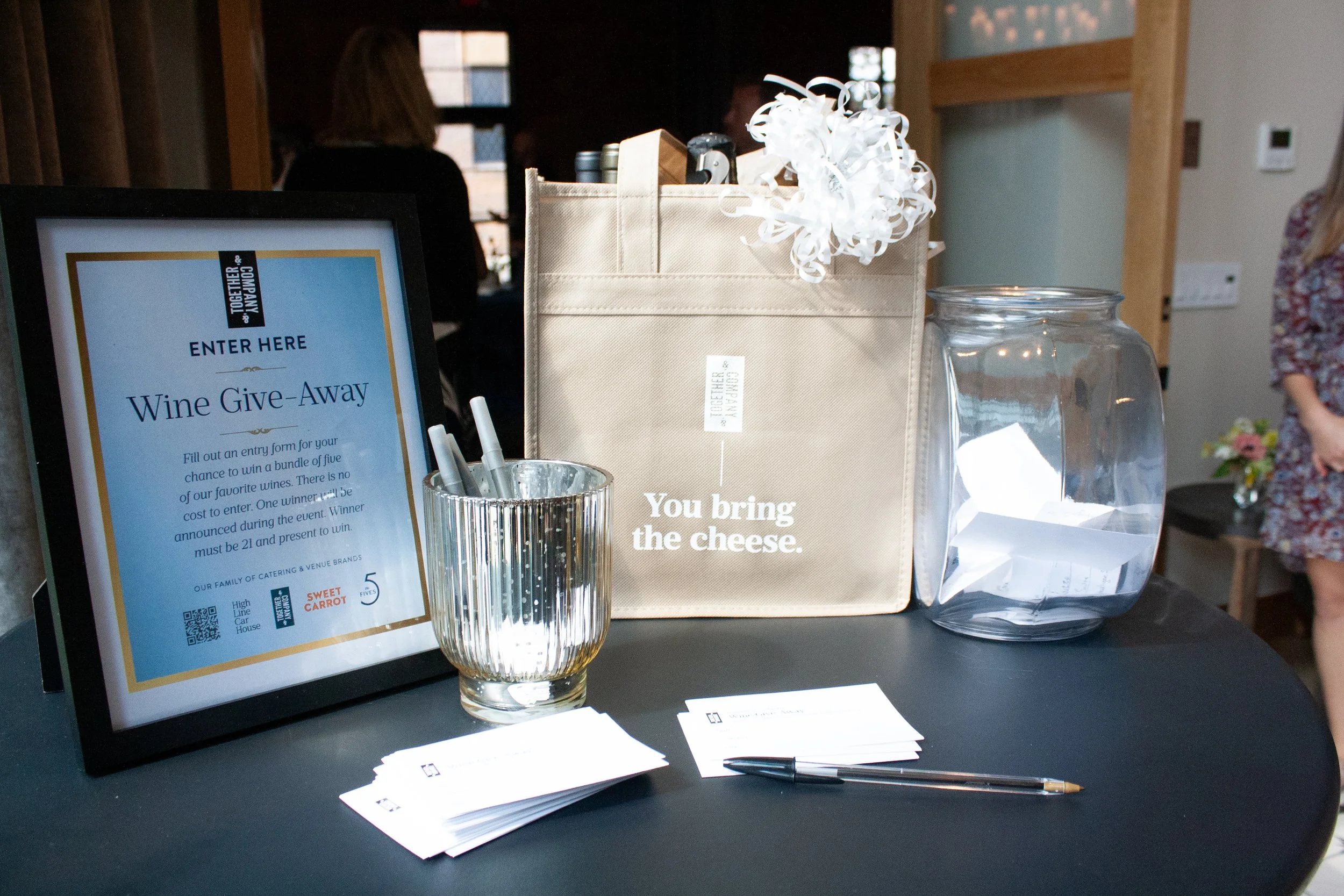 A wine give-away table at a corporate event in the grand entrance foyer at High Line Car House, including a sign, a gift bag, and a jar for guests to enter their names into the raffle. 