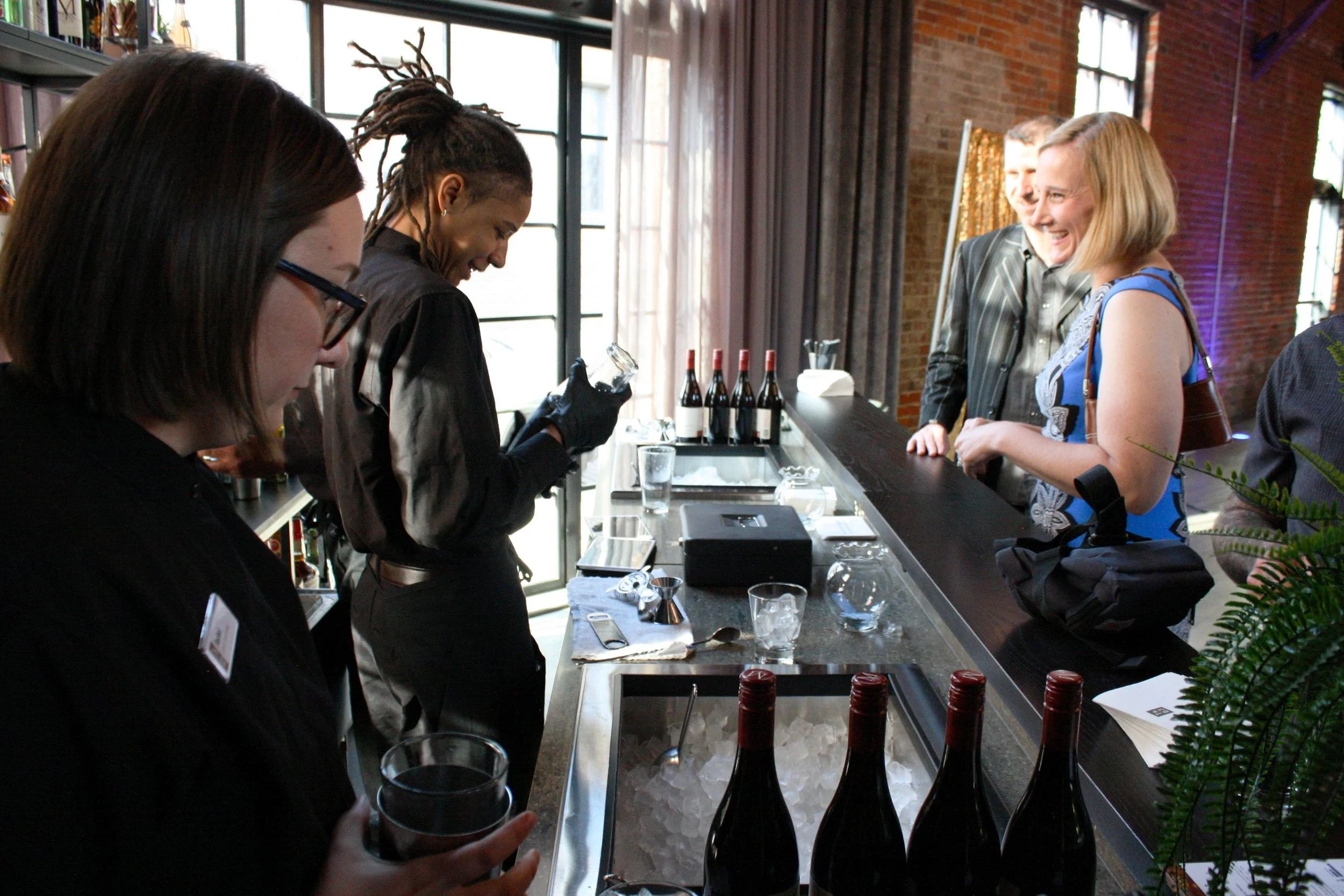 Guests at a fundraiser smile as they wait at the bar for a bartender to shake a cocktail for them.