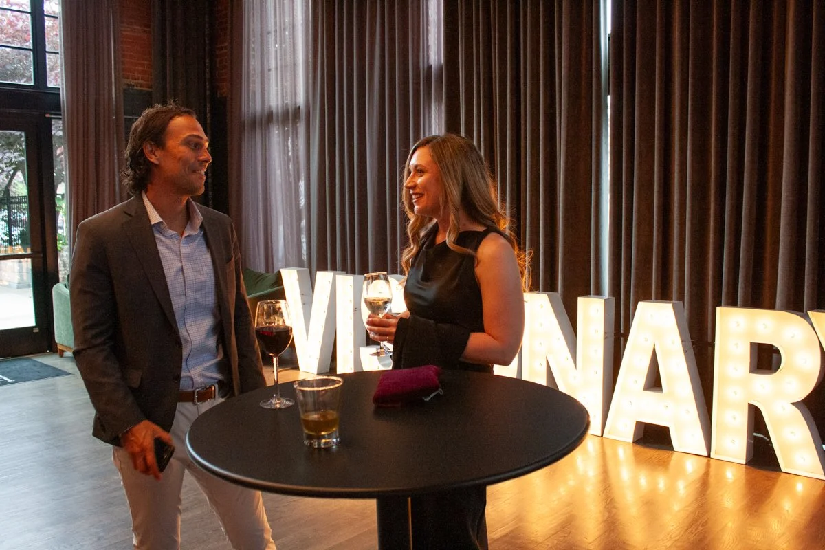 Two guests chat at a nonprofit fundraising event, holding glasses of wine and standing in front of large marquee letters that spell "Visionary". 