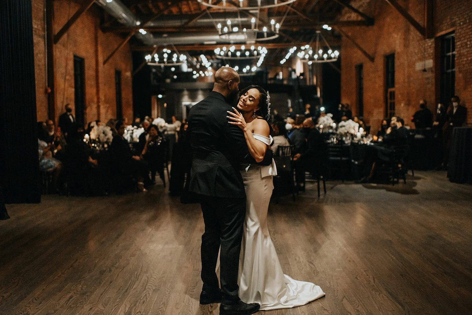 A newlywed couple share their first dance surrounded by warm brick walls and romantic two-tiered chandeliers.