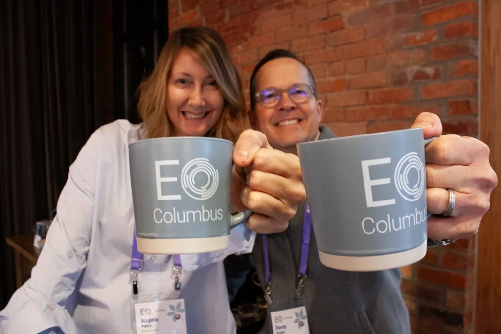 Two smiling colleagues hold branded coffee mugs towards the camera at a corporate breakfast event at High Line Car House in German Village, Columbus, Ohio.