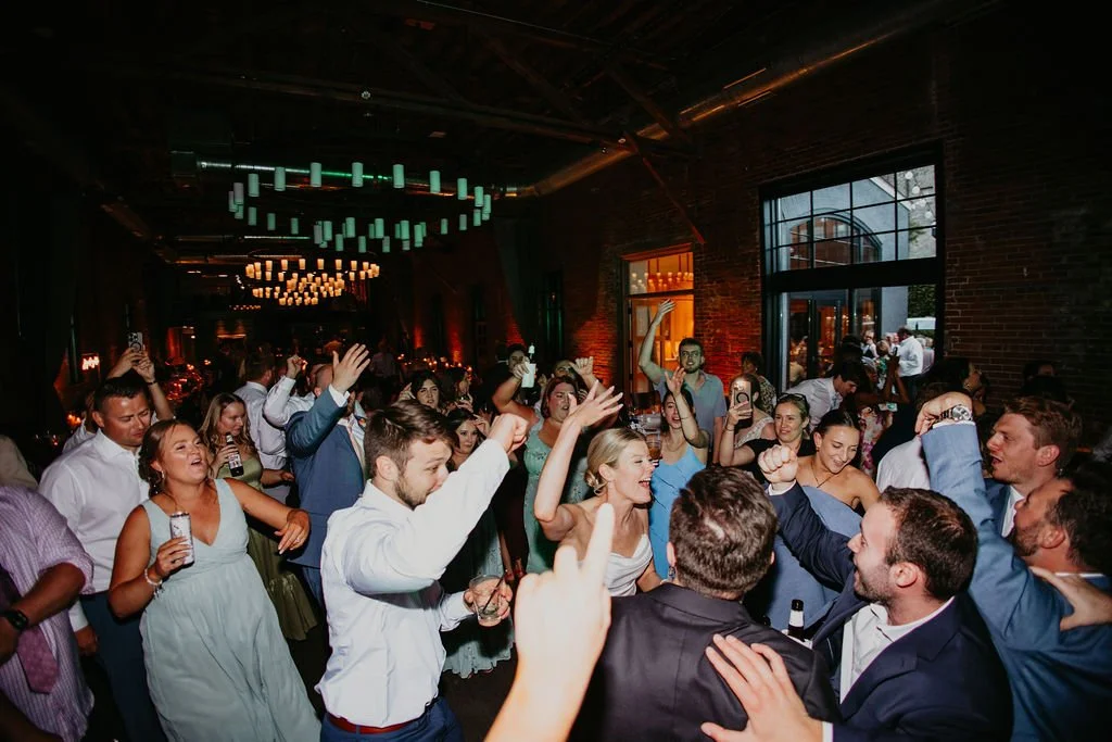 A bride leads the center of a dance floor at her reception. Many guests stand around her, singing with their hands up. The lights are dimmed in the reception hall.