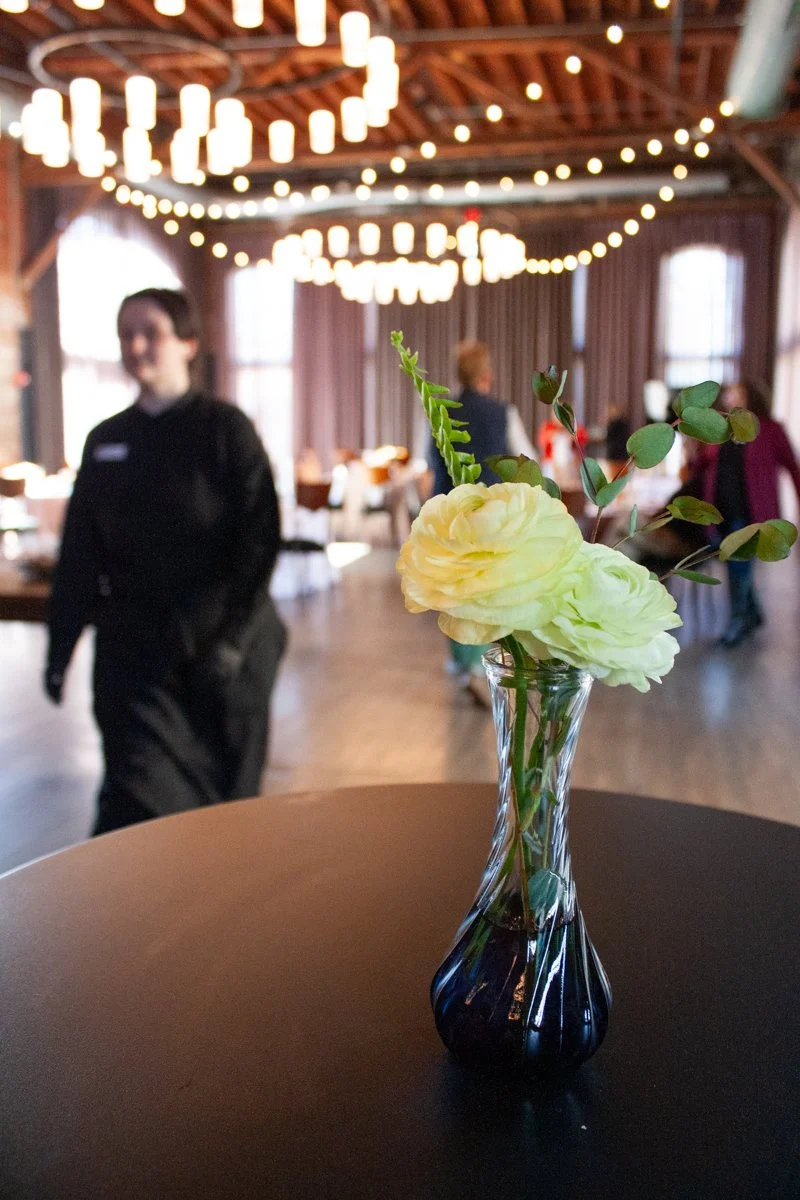 A votive floral centerpiece sits on a cocktail cab table in a spacious event space as a server from Together & Company catering walks by. 