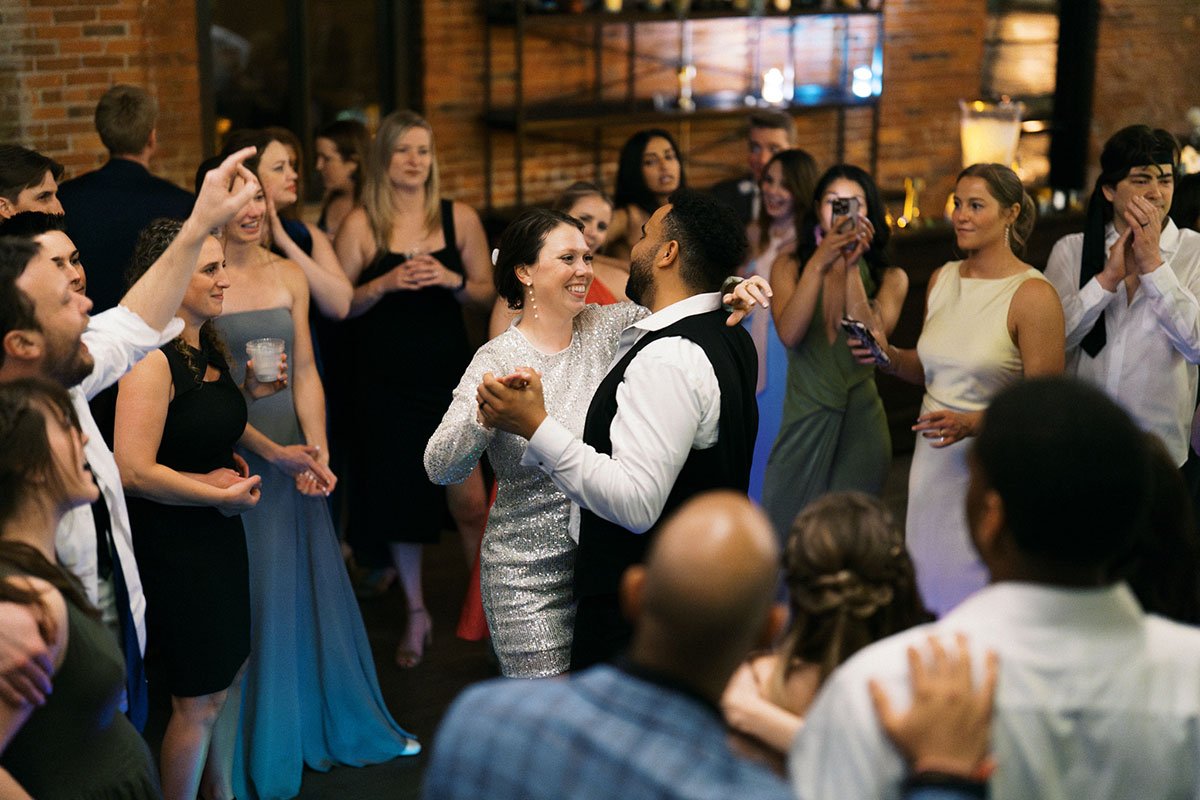 A bride and groom share a dance at their reception at a historic wedding venue in German Village, smiling at each other. The bride is wearing a silver sparkly dress. Their guests stand around them, watching and smiling.