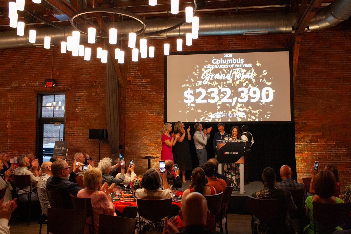 Guests at a nonprofit fundraising event applaud as a large monetary number shows up on a big screen above a stage.