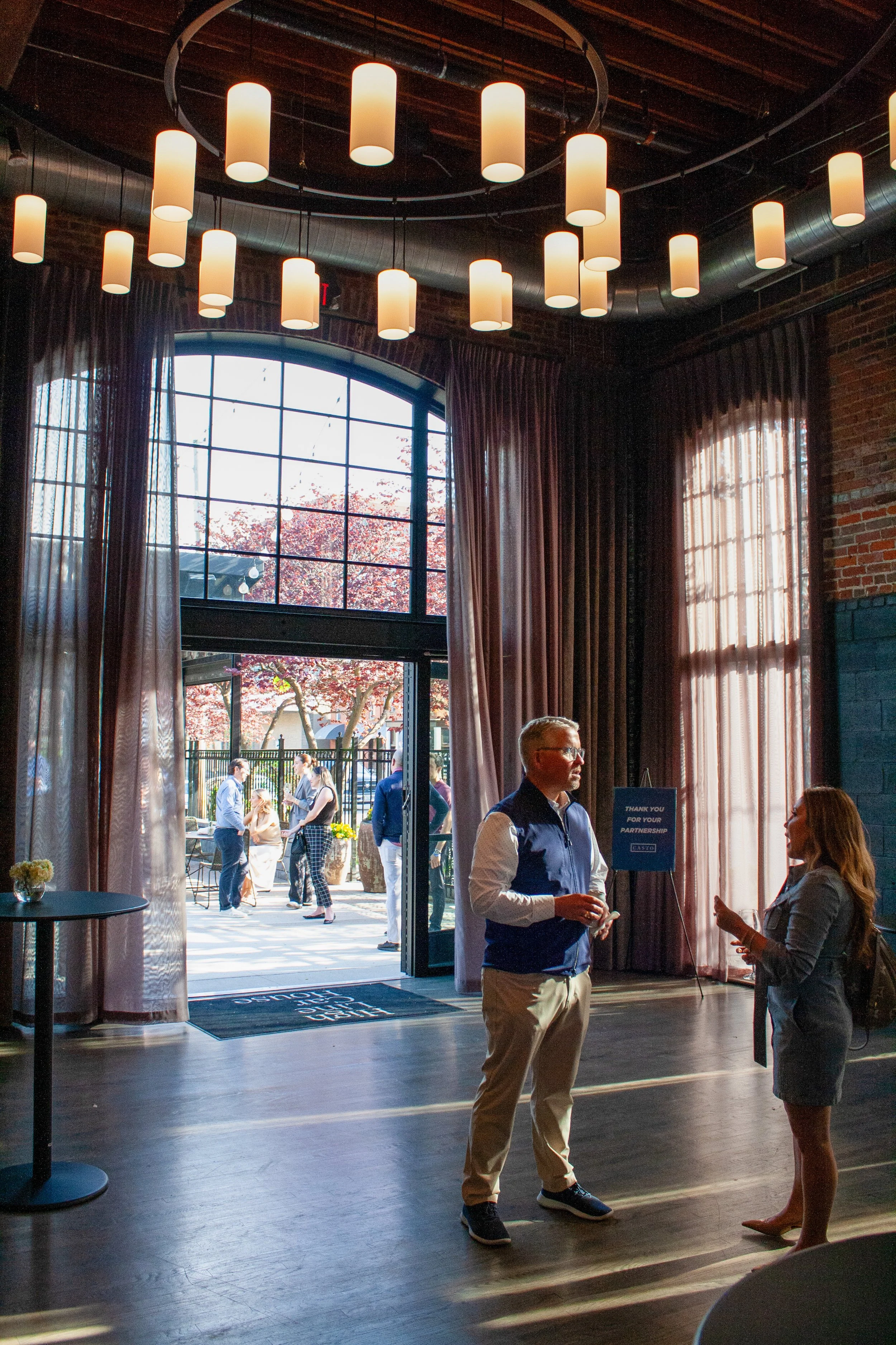 Two people talk business at a corporate happy hour in High Line Car House's grande hall. Behind them, the doors of the event space are open to the sunny outdoor patio, where other guests mingle. 