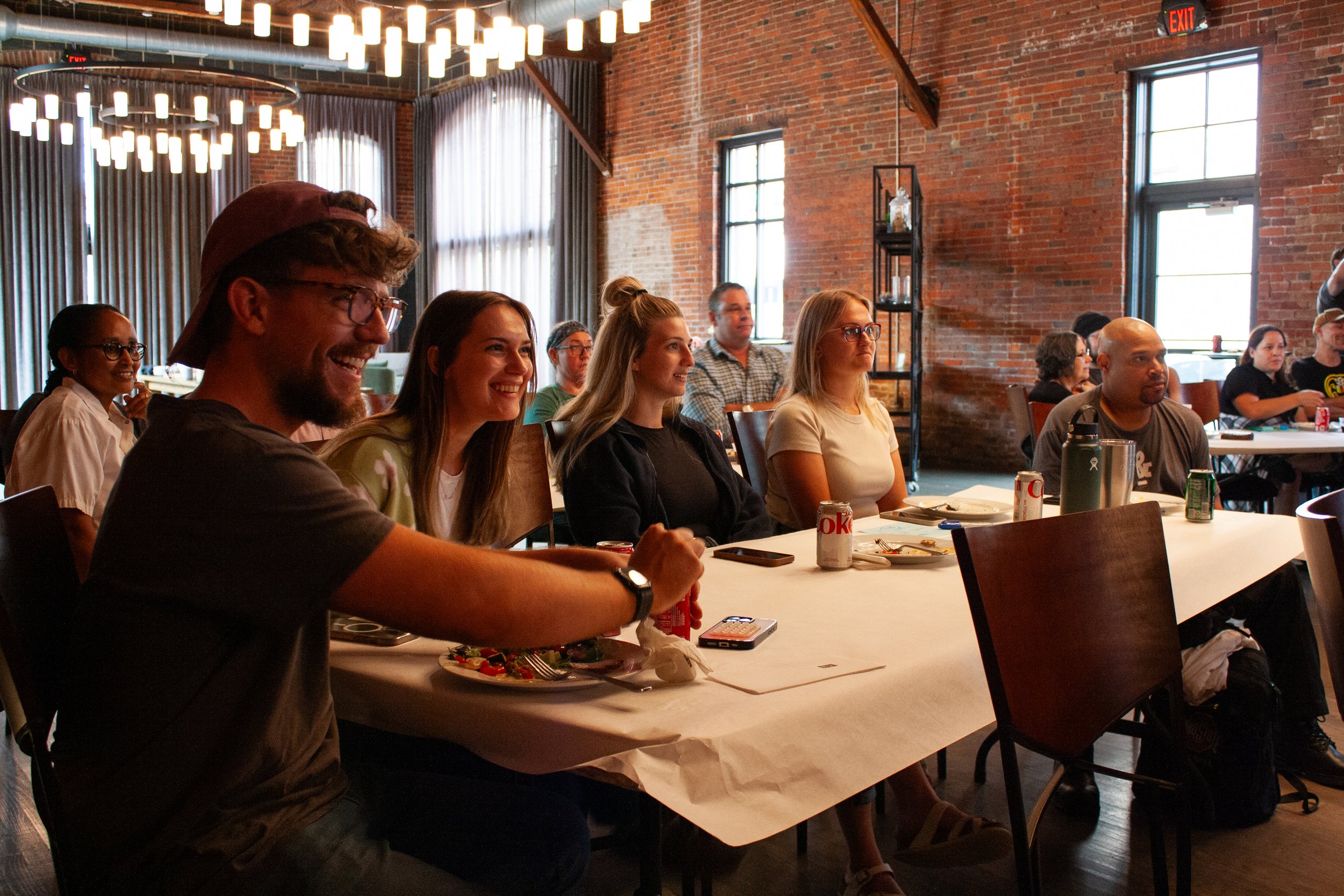 A group of coworkers sit together at a table during a company meeting in a large event space in Columbus, Ohio, laughing as they eat lunch and listen to the speaker, out of frame. 
