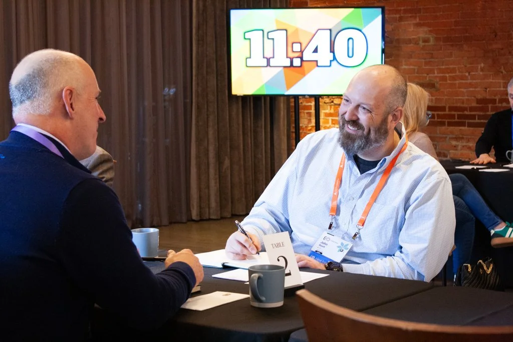 Two people sit at a table across from one another at a corporate gathering, one of them talking and the other taking notes. A timer ticks down on a screen behind them, next to floor-to-ceiling curtains that make the venue feel smaller. 