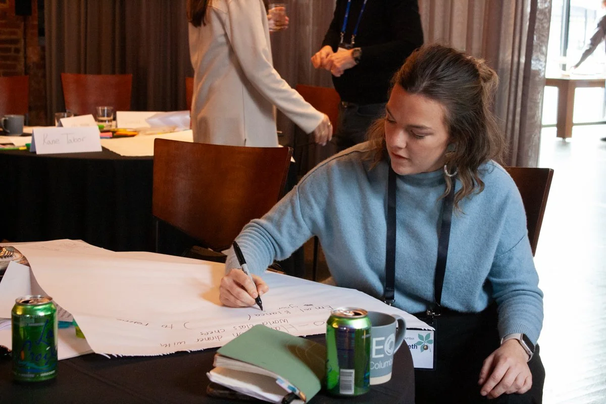 A corporate networking event attendee writes notes on a large post-it note. They're seated at a table in a large event space that's been divided with curtains to make it smaller. 