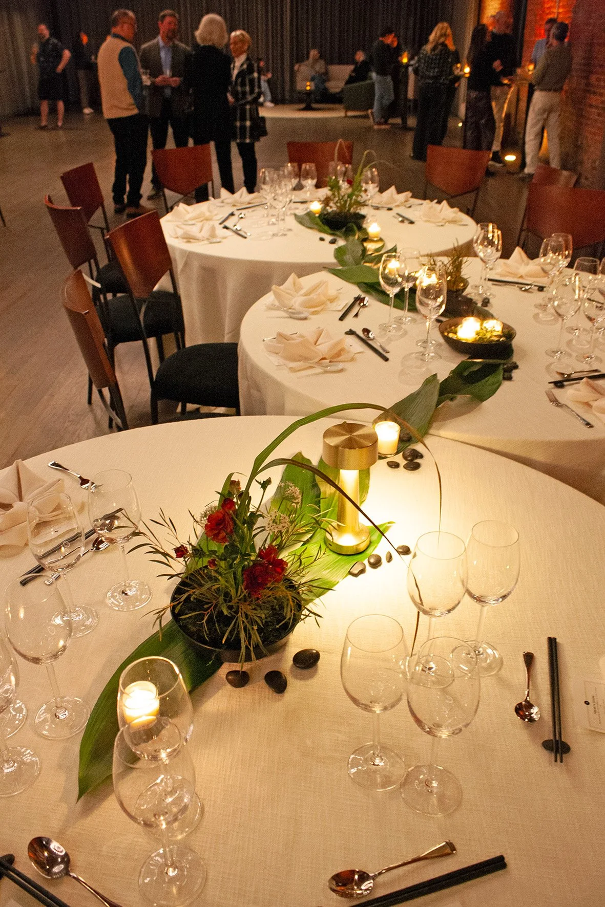 Long leaves and modern floral centerpieces are placed down the center of three round tables that are placed right next to each other in a winding shape. Votive candles are dotted along the way, creating a glow in the industrial event space.