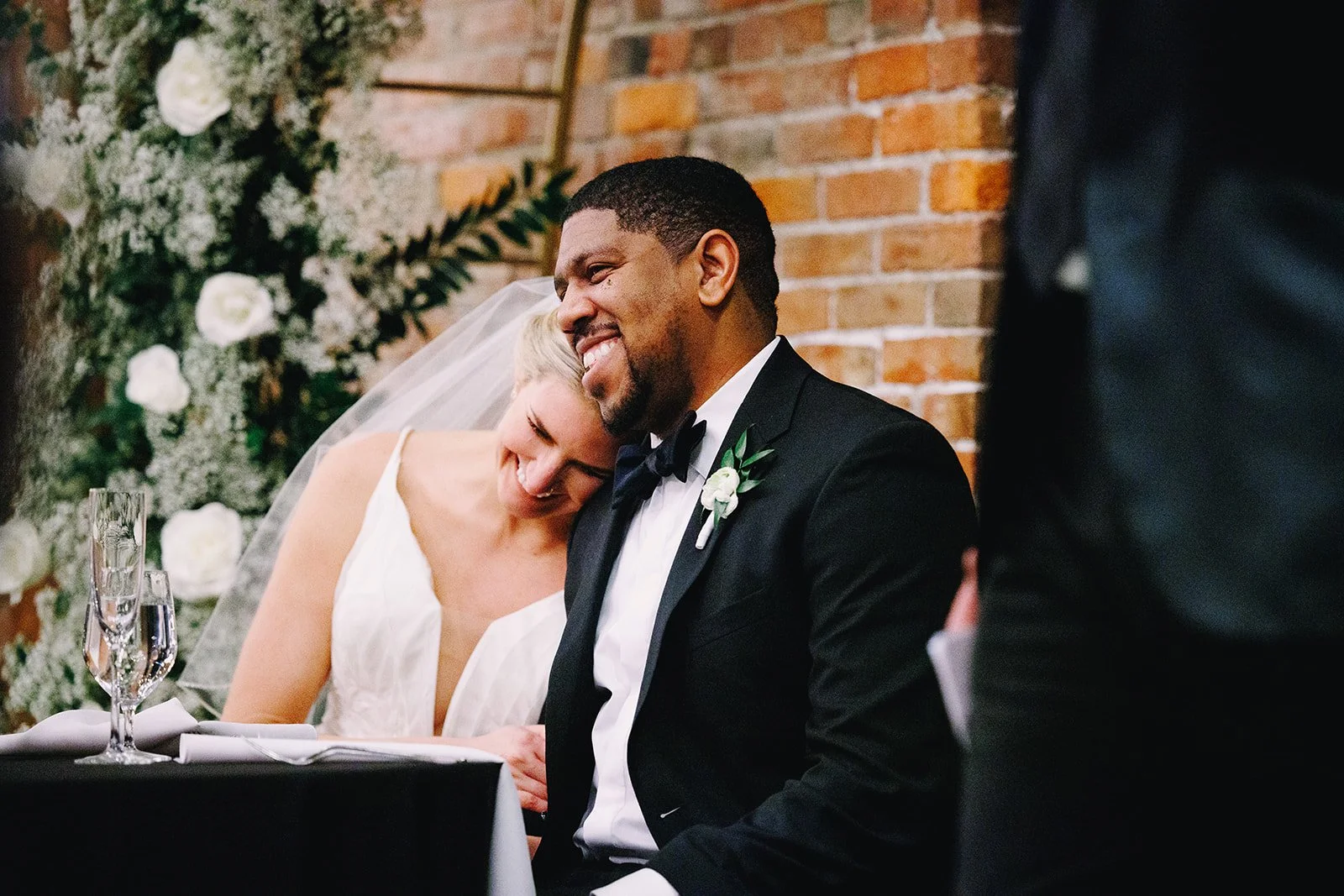 A bride leans on her groom's shoulder at their cozy and intimate wedding reception. Behind them, an arch of romantic white flowers against a warm brick wall frames the couple.