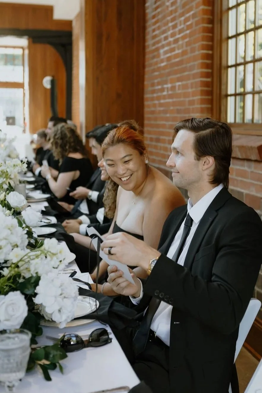 A wedding guest is seated at a long table and opens a note, smiling.