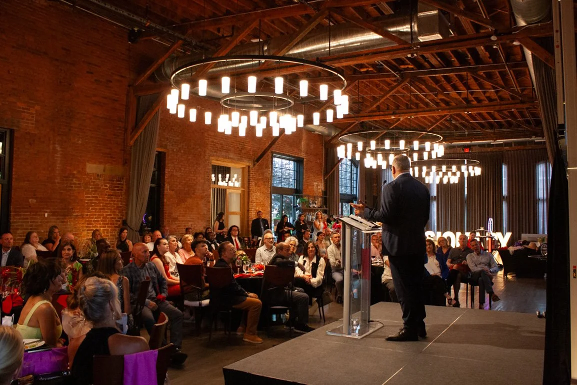 Someone stands on a stage with a podium and speaks to a crowd seated at round dinner tables in a large event space in Columbus, Ohio
