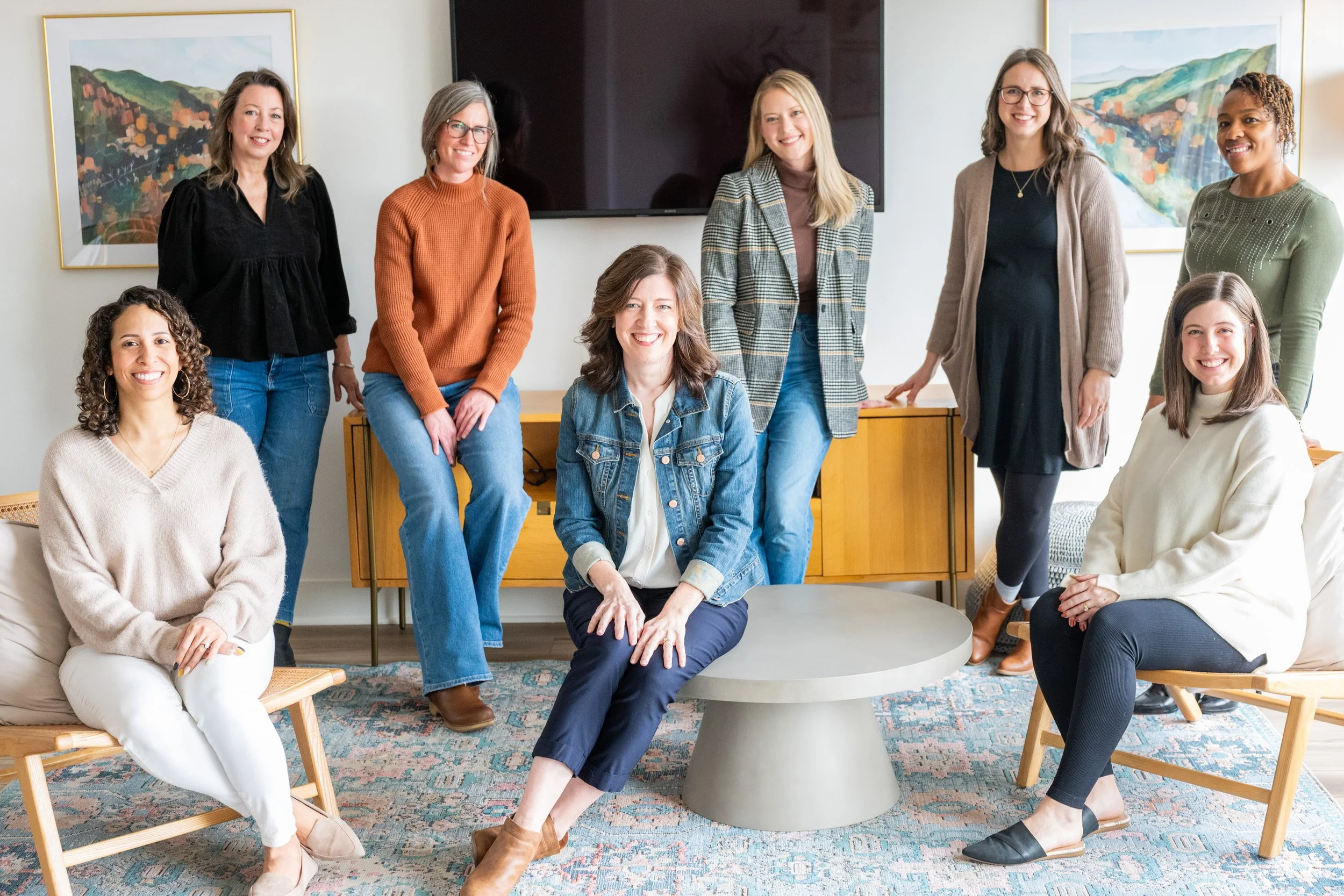 The team of All Belong Co, a short term rental management company based in Lynchburg, VA. A group of nine women of diverse ethnicities smiling and posing together in a living room with artwork on the walls and a television behind them.