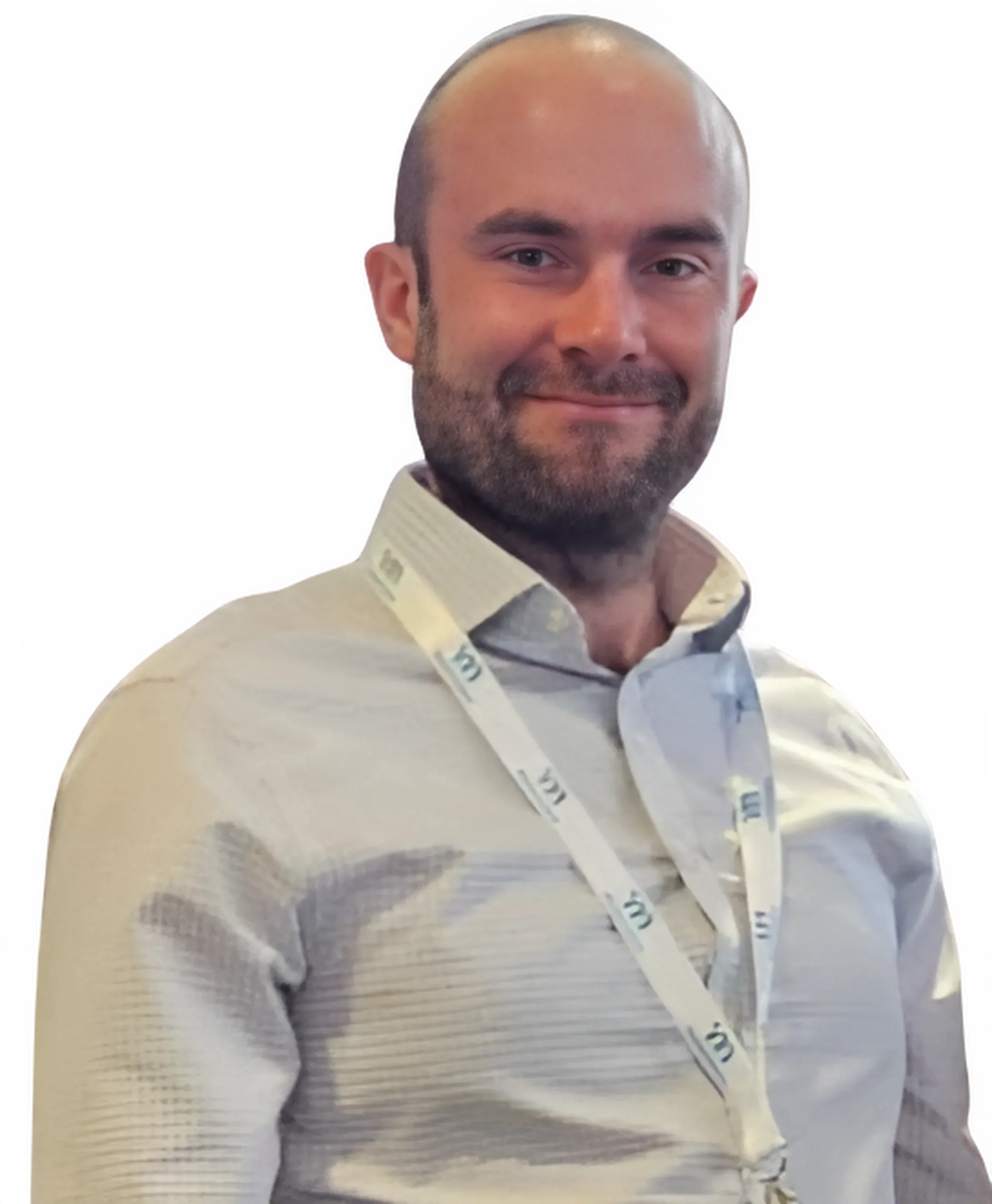 A man with a beard smiling, wearing a light-colored dress shirt and a conference badge around his neck, standing indoors.