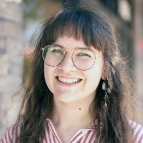 A smiling young woman with long brown hair and glasses, standing against a pastel-colored brick wall.