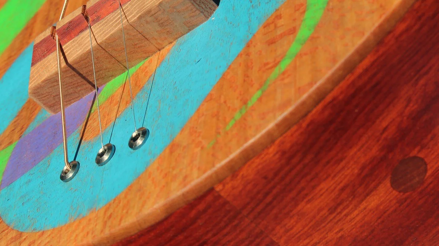 A three string acoustic/electric guitar with a snake motif inspired by the work and creativity of Freeman Vines. Built with padauk and sycamore woods, and painted with mineral paint. The head is carved in the shape of a forked tongue and the back fea