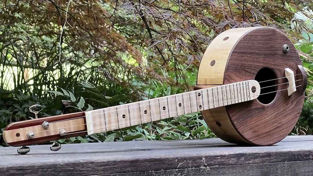This acoustic/electric guitar is built with a circle theme. The strings are tuned to D, A, E. The woods include tiger maple, walnut and padauk. The back is painted with mineral paint and distressed. A circle starburst can be seen inside the sound hol