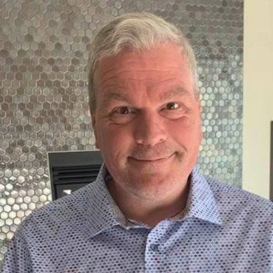 A middle-aged man with gray hair and a light beard smiling, wearing a light blue patterned shirt, standing in front of a metallic hexagonal tiled wall.
