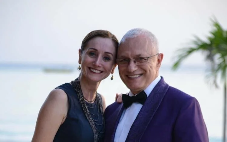 A smiling woman in a navy dress and a smiling man in a tuxedo stand close together outdoors near water with a beach and palm tree in the background.