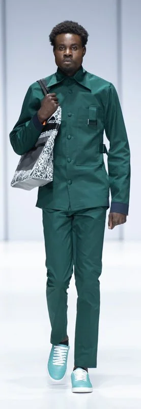 Model walking on a runway wearing a green outfit with a matching button-up shirt and pants, teal sneakers, and carrying a patterned tote bag.