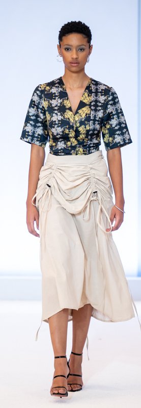 A fashion model wearing a floral blouse and a draped, beige midi skirt with strappy high heels on a runway.