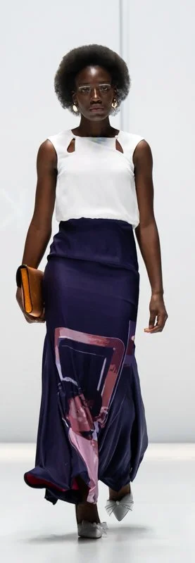 Model walking on a runway wearing a white sleeveless top and a long, printed navy skirt, holding an orange clutch.