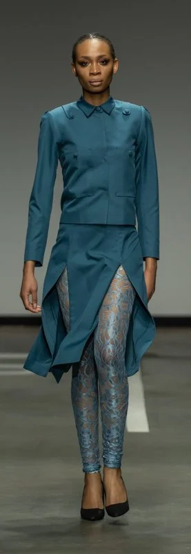 Model wearing a teal jacket and skirt with patterned leggings and black shoes on a runway.