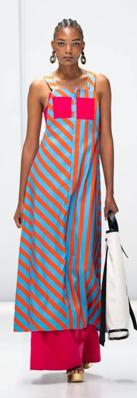 Model wearing a colorful striped dress with blue and orange pattern, holding a white bag, on a runway.